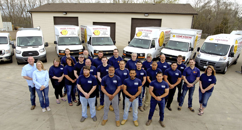 Duane Blanton Plumbing staff standing together in parking lot with row of parked service trucks behind them.